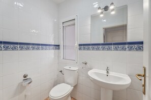 Un baño con azulejos blancos, un inodoro, un lavamanos y una ventana.