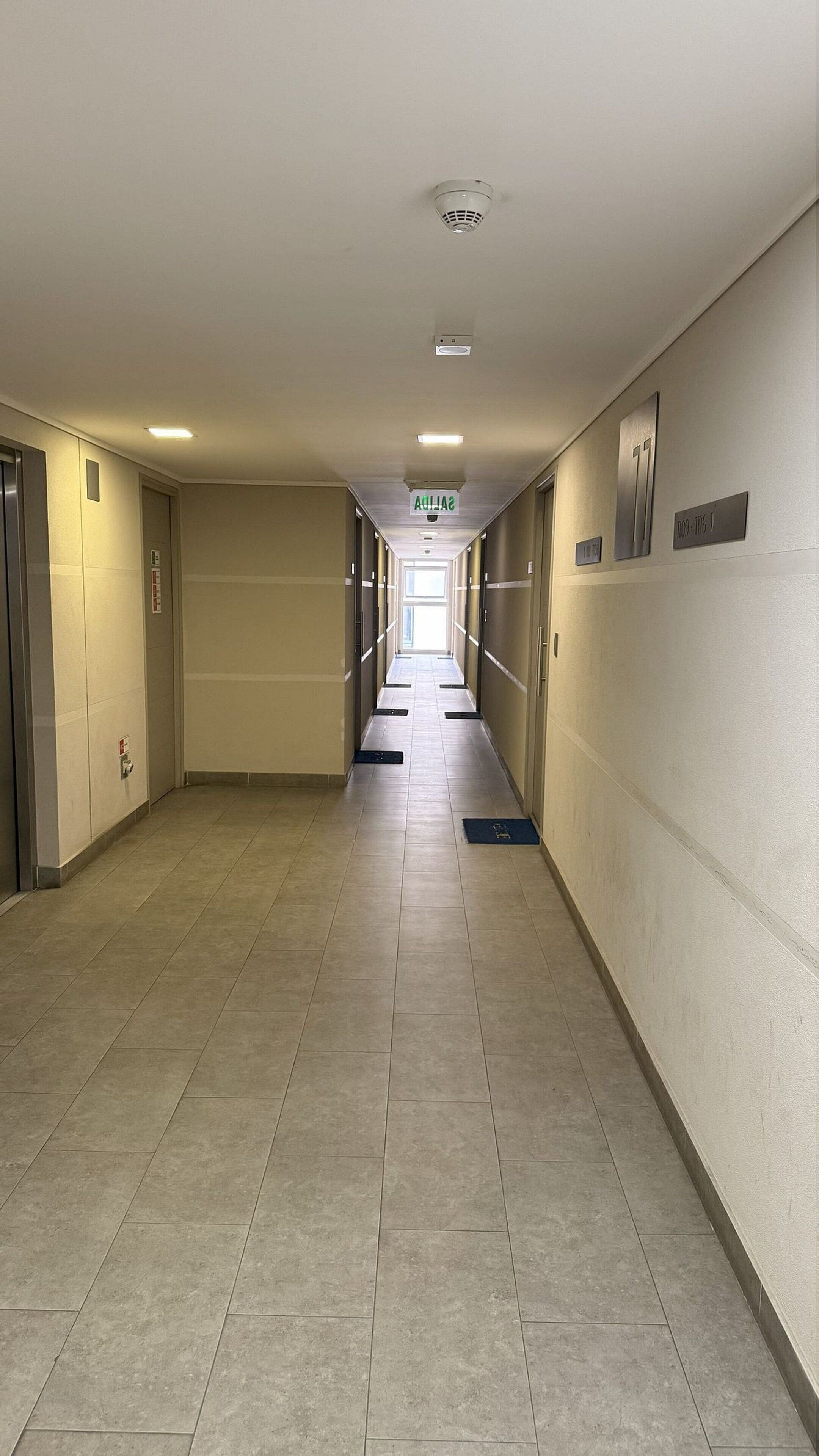 Un largo pasillo de hotel estrecho, con suelo de baldosas, puertas a ambos lados y un letrero que indica la dirección al ascensor.
