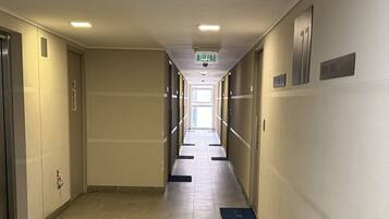 Un largo pasillo de hotel estrecho, con suelo de baldosas, puertas a ambos lados y un letrero que indica la dirección al ascensor.