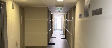 A long, narrow hotel corridor with tiled floors, doors on both sides, and a sign indicating the direction to the elevator.