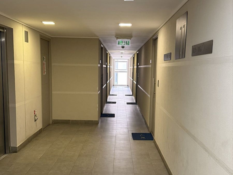 Un largo pasillo de hotel estrecho, con suelo de baldosas, puertas a ambos lados y un letrero que indica la dirección al ascensor.