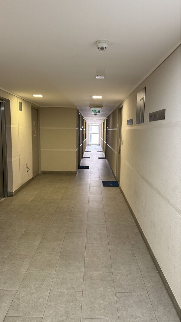 A long, narrow hotel corridor with tiled floors, doors on both sides, and a sign indicating the direction to the elevator.