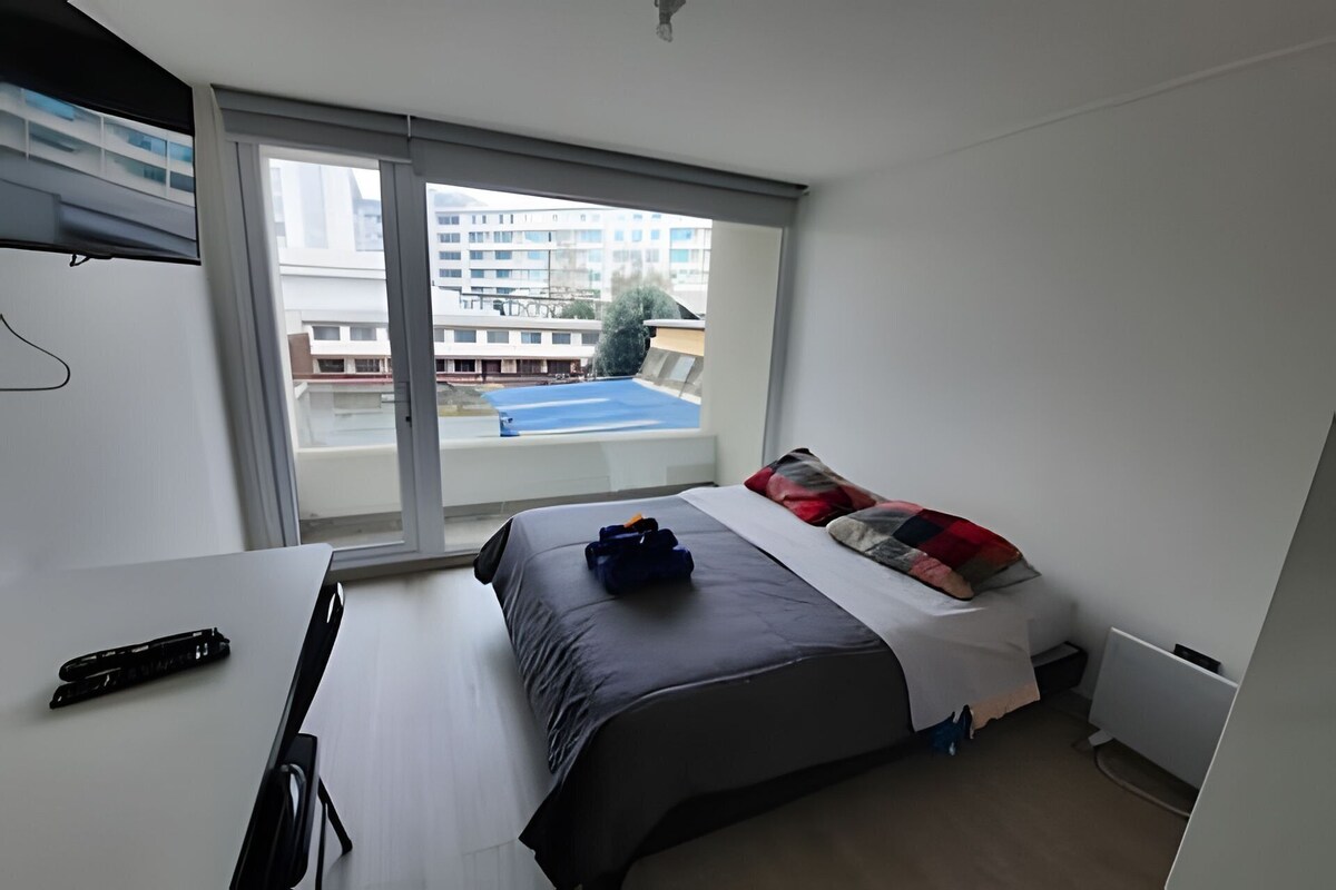 A bedroom with a bed, a desk, and a window overlooking a pool and buildings.
