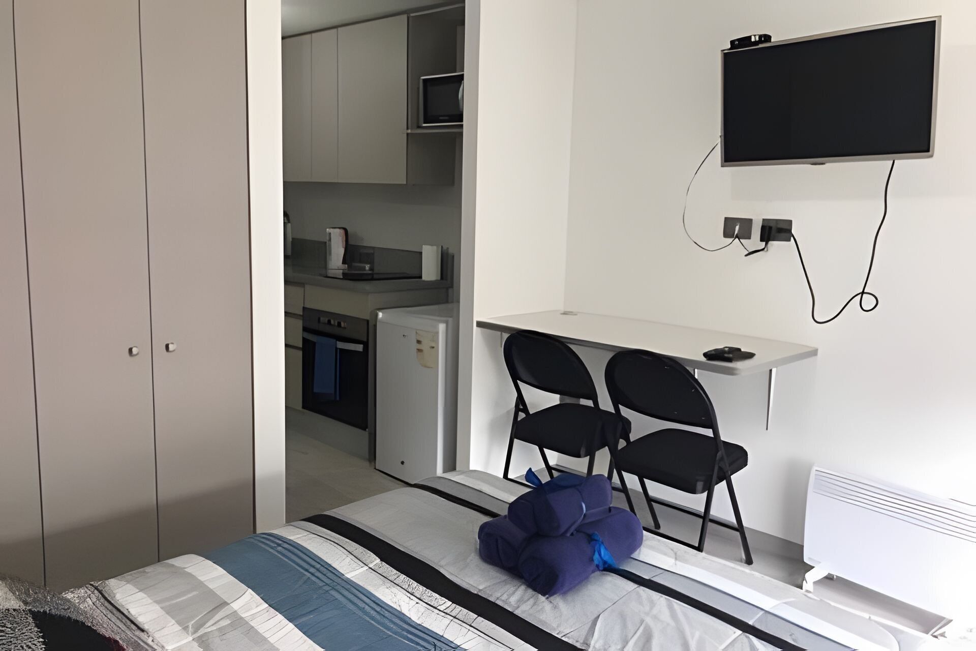 Une chambre à coucher avec un lit, un bureau avec deux chaises, une télévision et, au fond, une petite cuisine.
