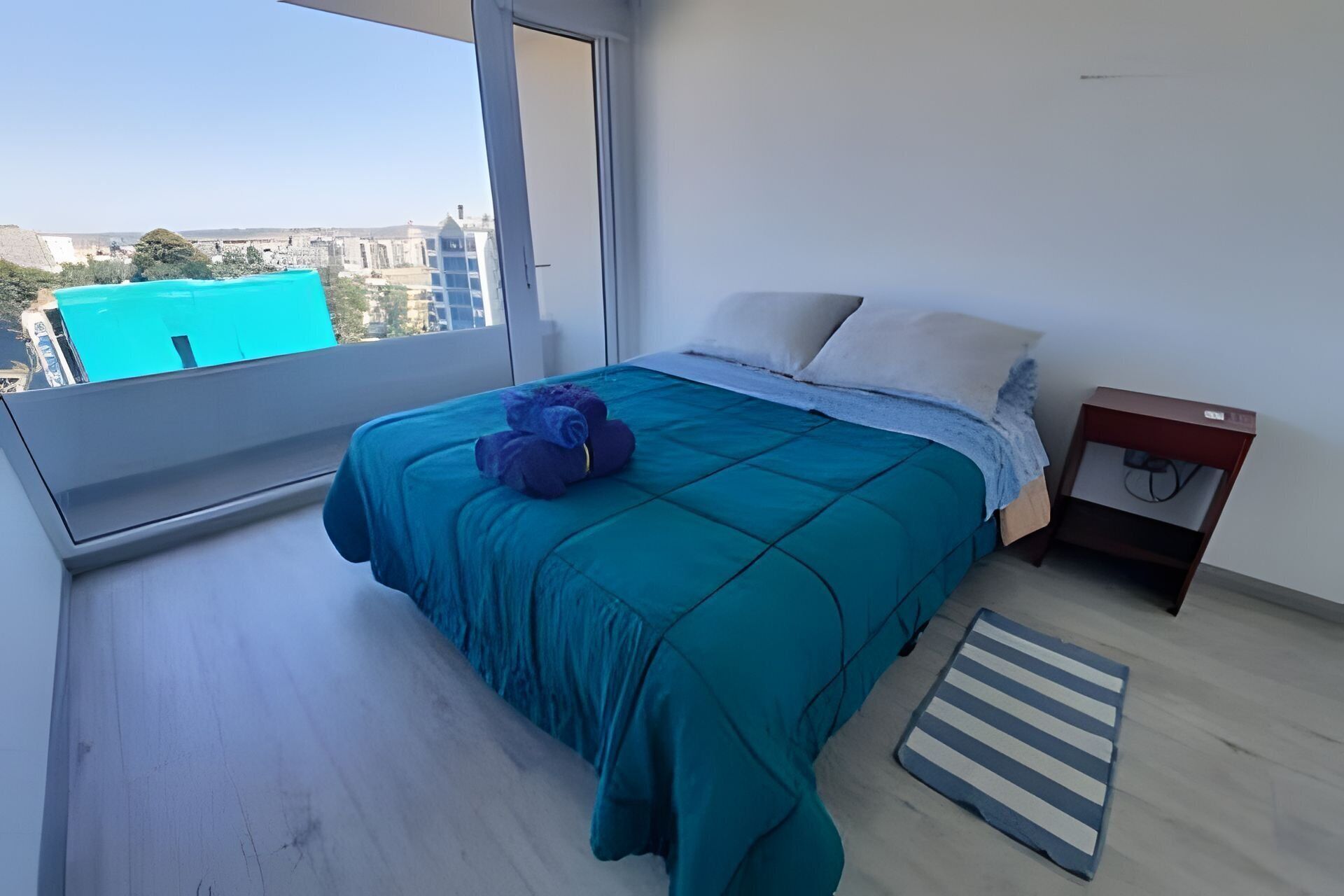 Une chambre avec une grande fenêtre, un lit avec une literie de couleur turquoise, une table de chevet et une vue sur des immeubles.