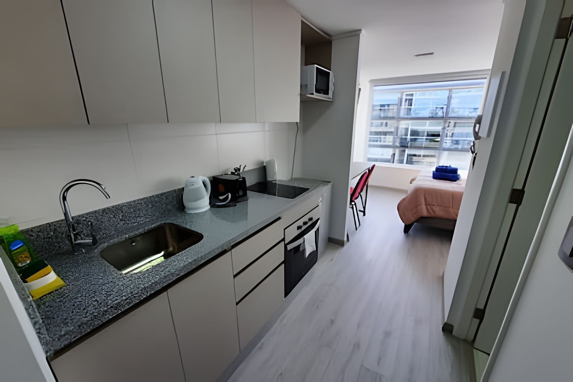 Una cocina moderna con encimera de granito, pileta de acero inoxidable y microondas. Hay un pequeño comedor con una silla y una cama con una manta rosa.
