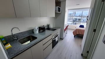 Cozinha moderna com bancada de granito, pia de aço inoxidável e micro-ondas. Há uma pequena área de refeições com uma cadeira e uma cama com manta rosa.