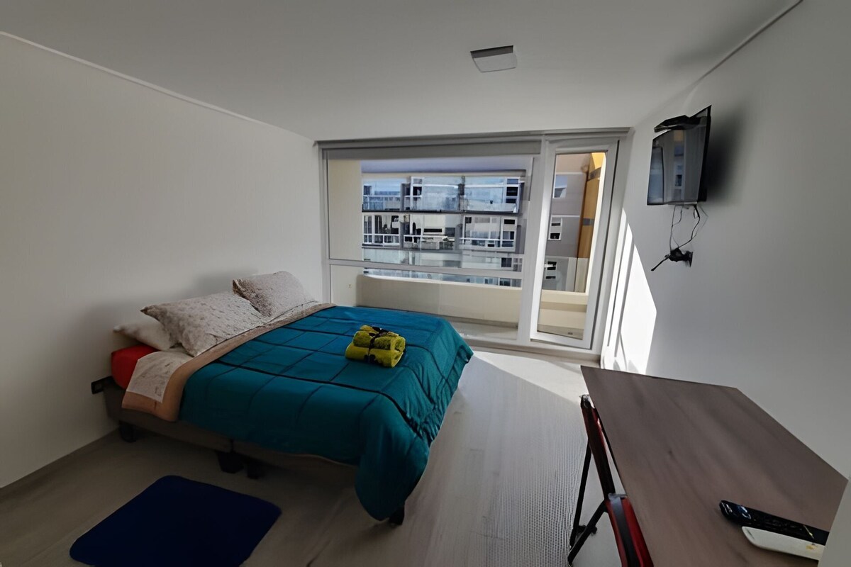 A bedroom with a bed, a desk, and a window overlooking a balcony.