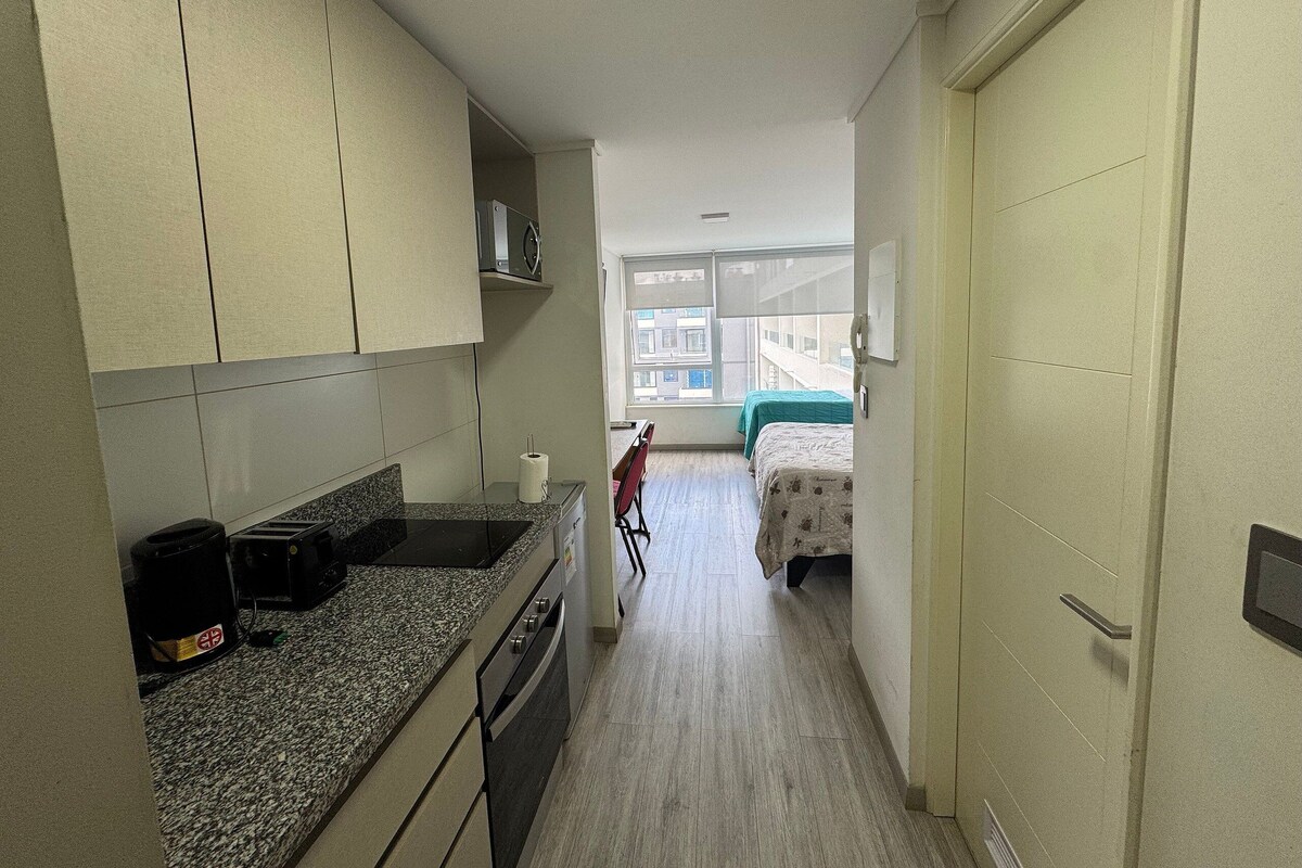 A narrow hallway with a kitchenette on one side, a bed with a green cover in the distance, and a window with a view of buildings outside.