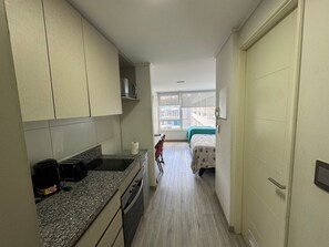 A narrow hallway with a kitchenette on one side, a bed with a green cover in the distance, and a window with a view of buildings outside. - Edificio Stepke (Temuco)