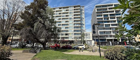 A multi-story residential building with balconies, a parking area with cars, and a small park with trees and a bench.