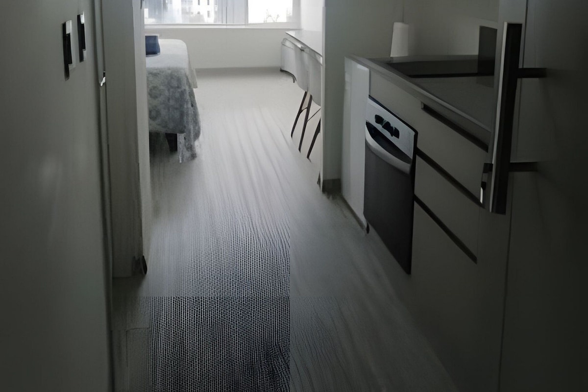 A modern hotel room with a kitchenette, a large window with a city view, and a neatly arranged bed.