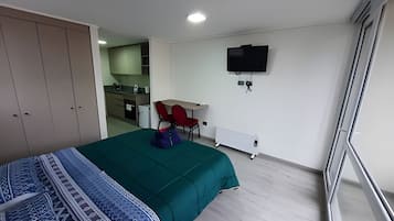 Un dormitorio con cama, escritorio, silla, televisor y una kitchenette al fondo.