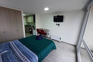 A bedroom with a bed, a desk, a chair, a TV, and a kitchenette in the background. - Edificio Stepke (Temuco)