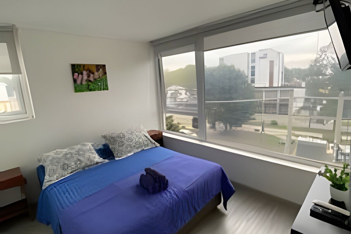 A bedroom with a bed, a desk, and a window with a view of buildings and trees.