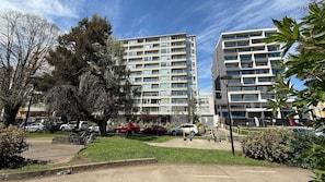 Un edificio residencial de varios pisos con balcones, un estacionamiento con automóviles y un pequeño parque con árboles y un banco.