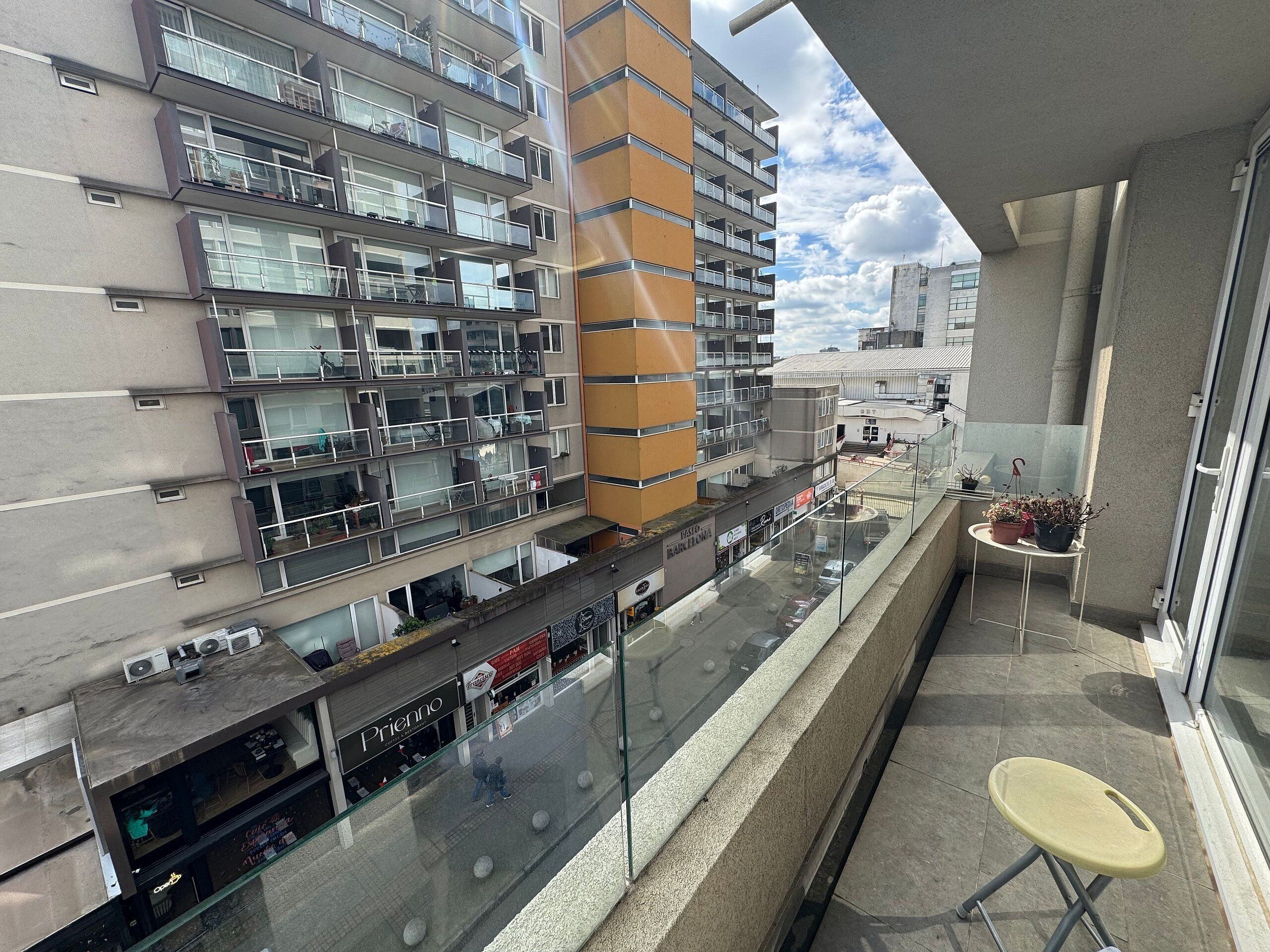 Un edificio de gran altura con varios balcones, barandilla de cristal y una pequeña mesa con plantas en macetas.