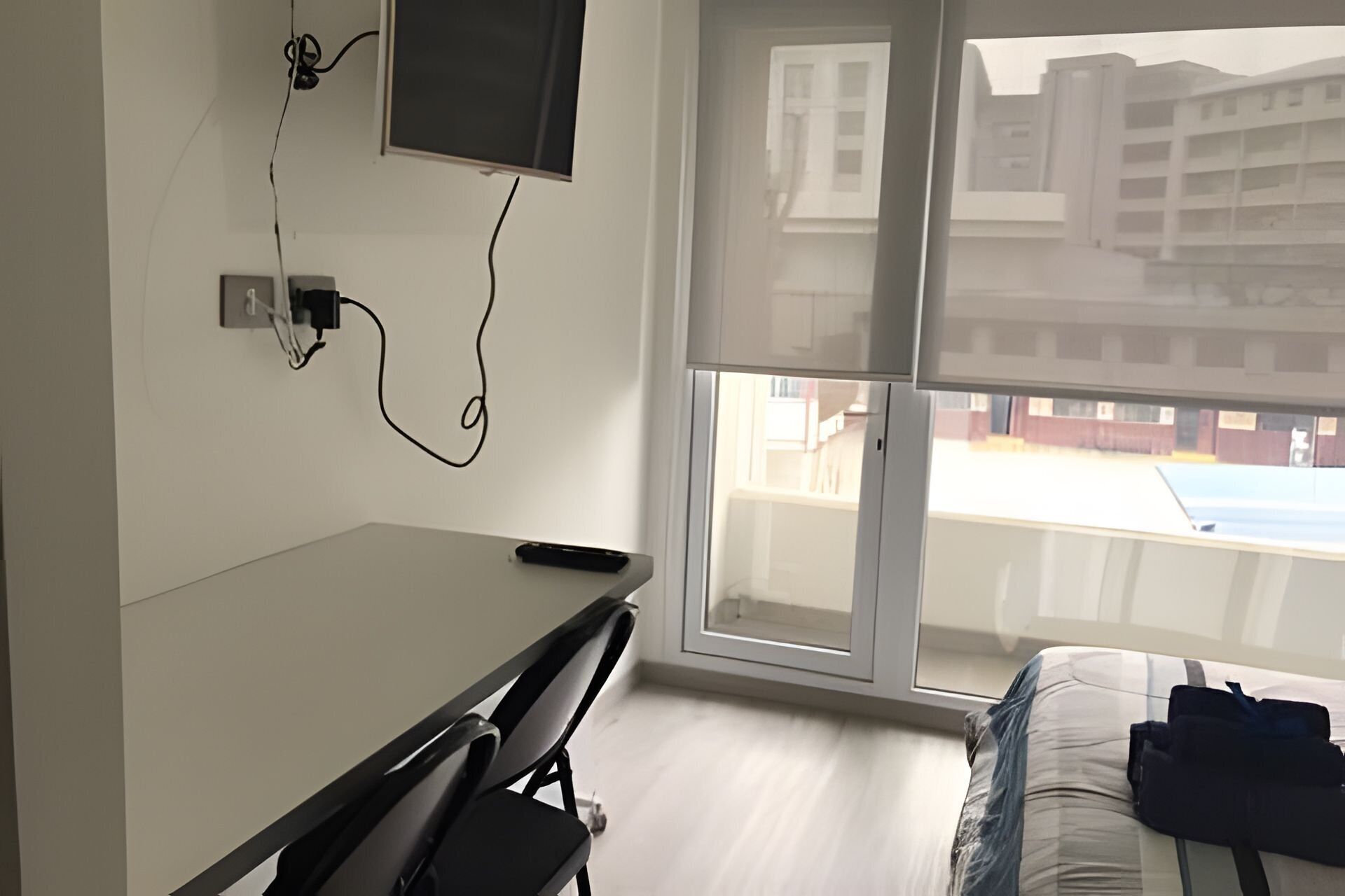 A hotel room with a bed, a desk, a chair, a TV mounted on the wall, and a large window offering a view of buildings outside.
