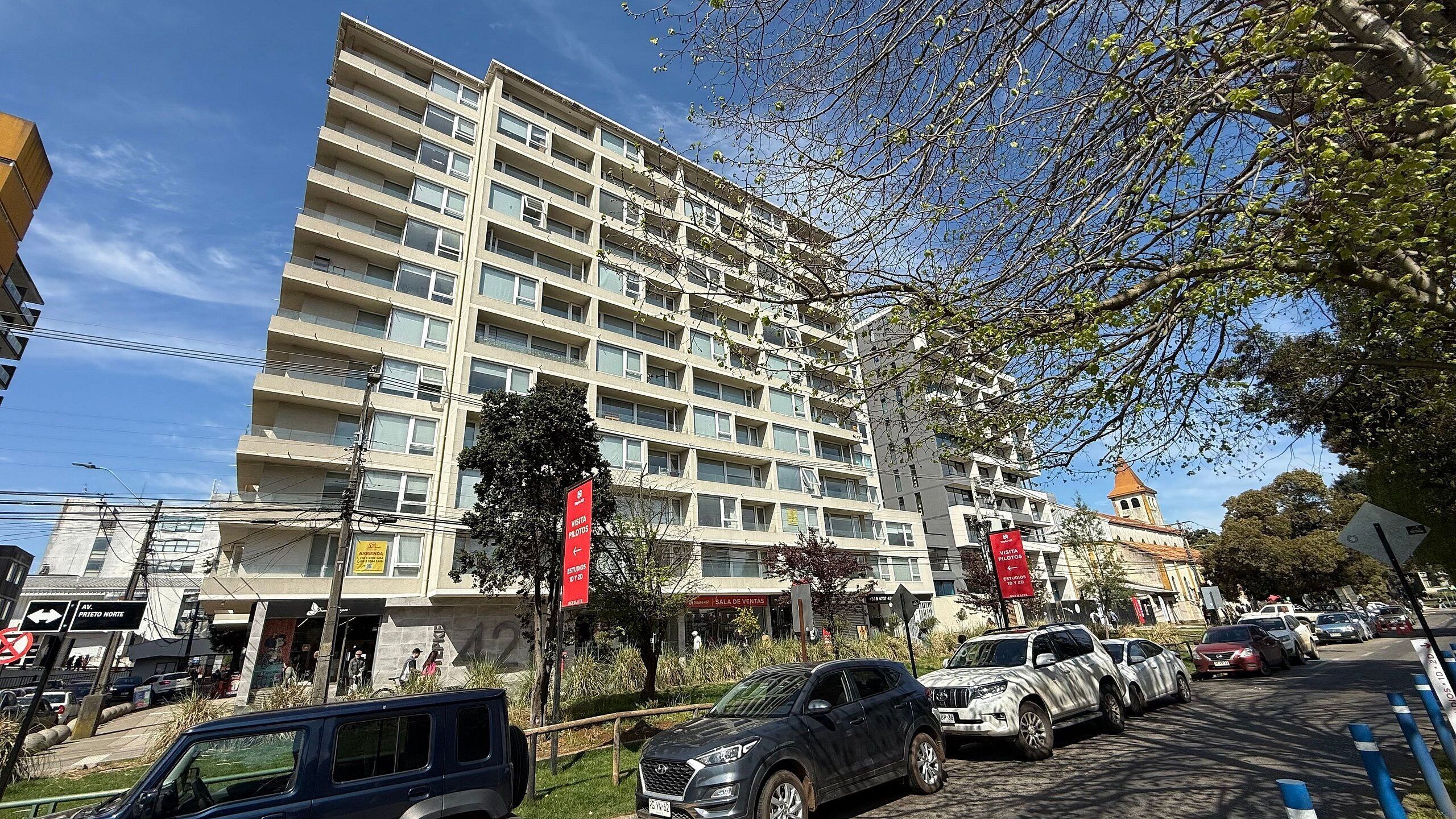 Un edificio de varios pisos con balcones, un estacionamiento con automóviles y un letrero rojo que indica una sala de ventas.