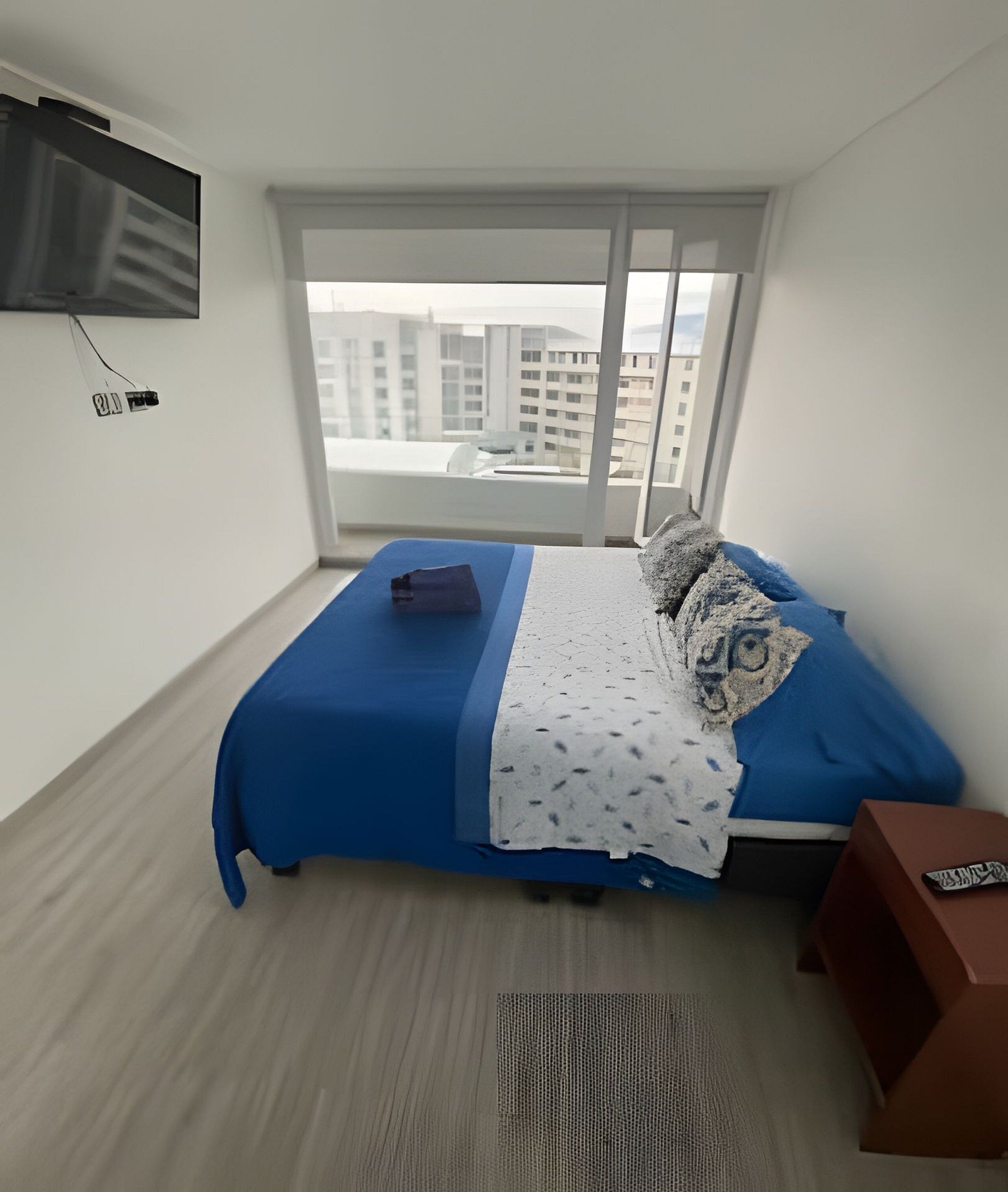 Un dormitorio con cama, televisor montado en la pared, ventana con vista a edificios y una mesita con control remoto.
