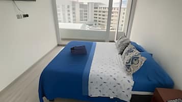 Un dormitorio con cama, televisor montado en la pared, ventana con vista a edificios y una mesita con control remoto.