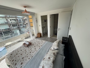 A bedroom with a bed, a small wooden stool, a remote control, and a view of a balcony with a railing.