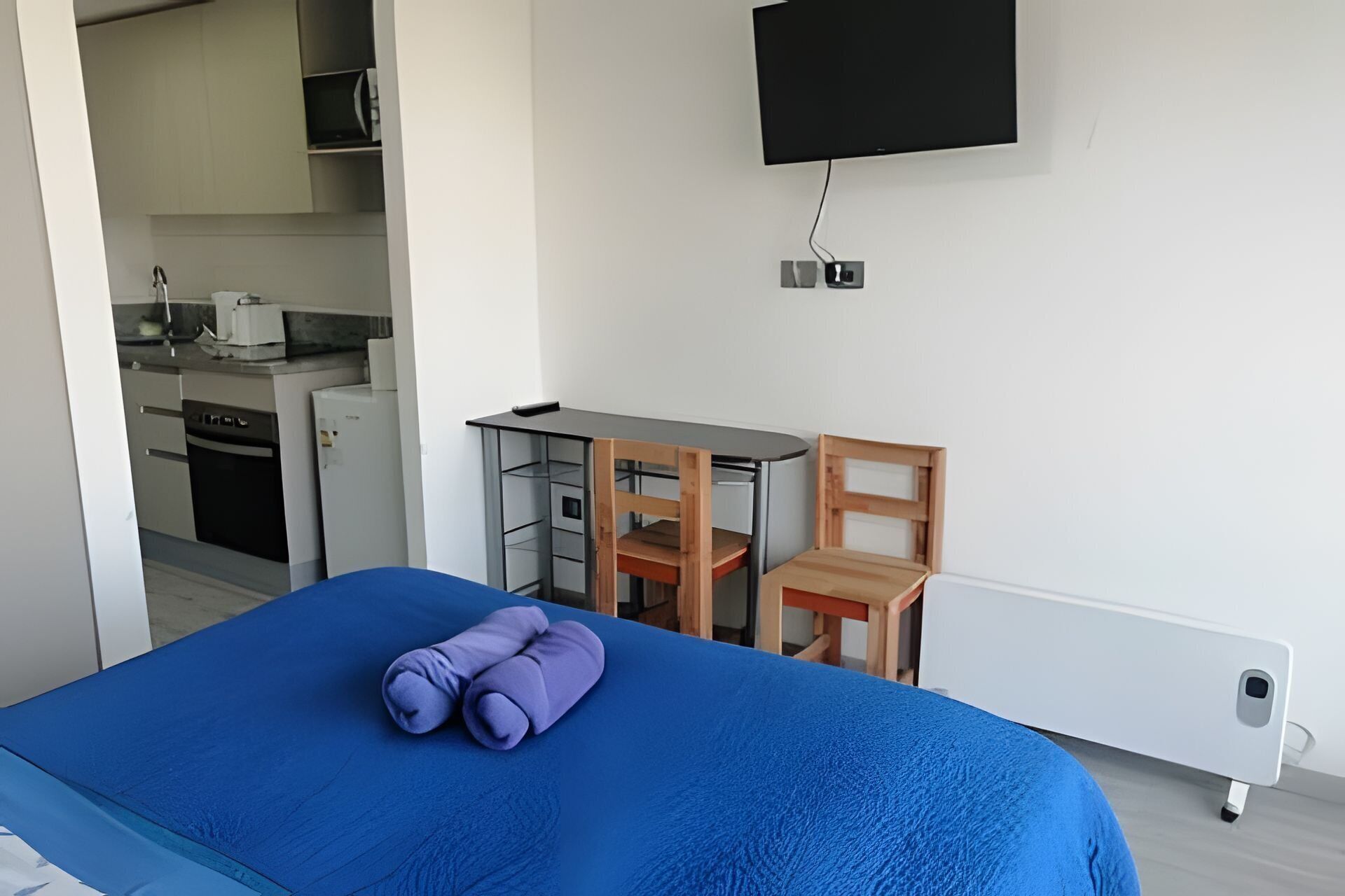Un dormitorio con cama, escritorio, silla, televisor y una kitchenette al fondo.