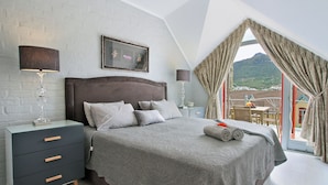Une chambre à coucher avec un grand lit, une table de chevet, une lampe, un tableau encadré et une vue sur l’extérieur.