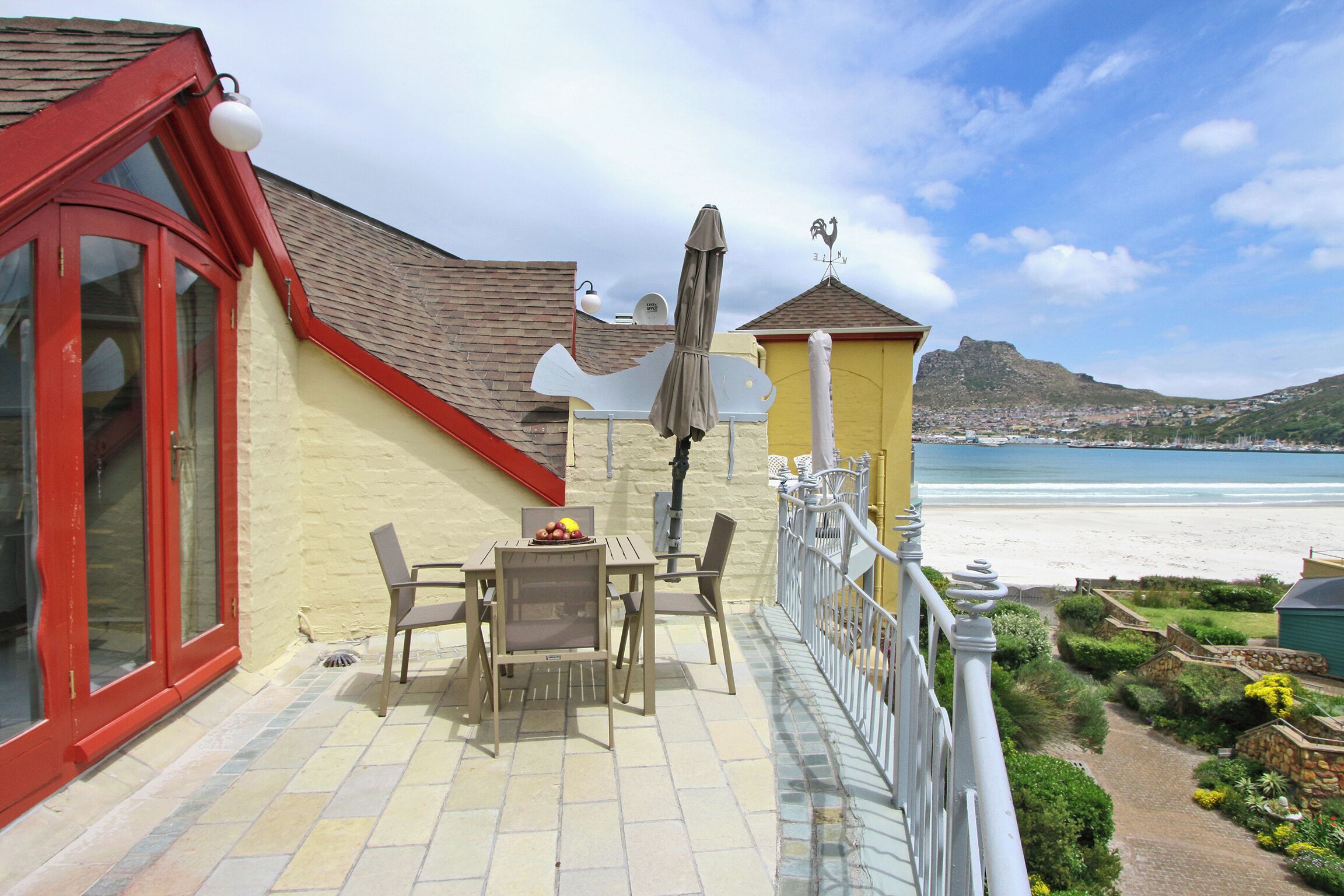 A balcony with a table and chairs, overlooking a beach and a mountainous landscape.