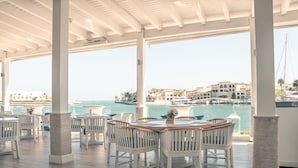 A covered outdoor seating area with white chairs and tables, overlooking a body of water and buildings in the background. - Studio 3 Guests Balcony With Views Fishing Lodge (Punta Cana)