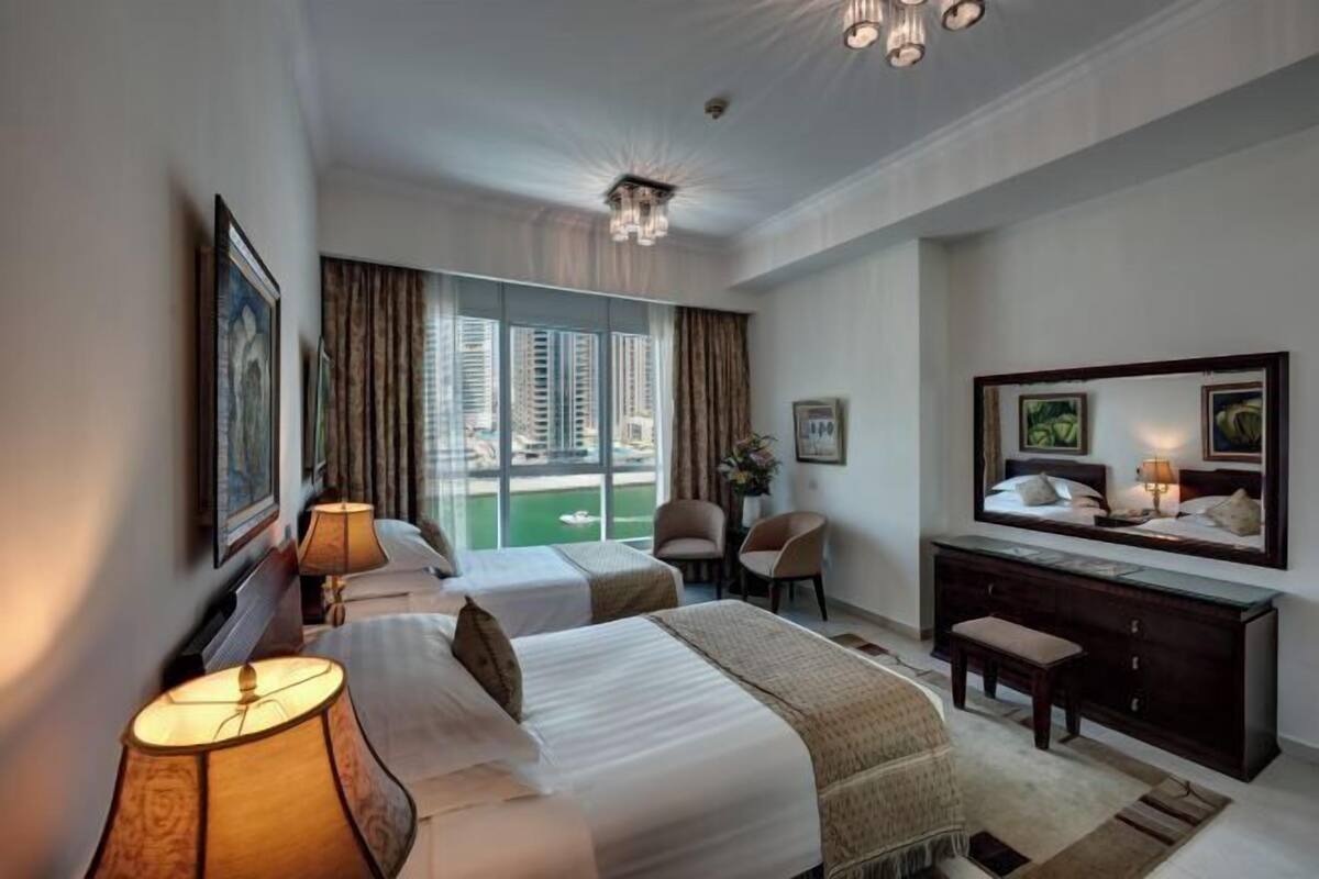 Une chambre d’hôtel avec deux lits, un bureau, une chaise, une commode et une grande fenêtre donnant sur des immeubles.