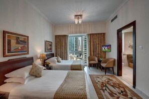 Habitación de hotel con dos camas, una televisión, un balcón con vistas a edificios y un comedor.