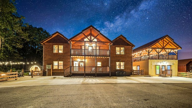 A two-story log cabin with a balcony, surrounded by a well-lit driveway and a starry night sky.