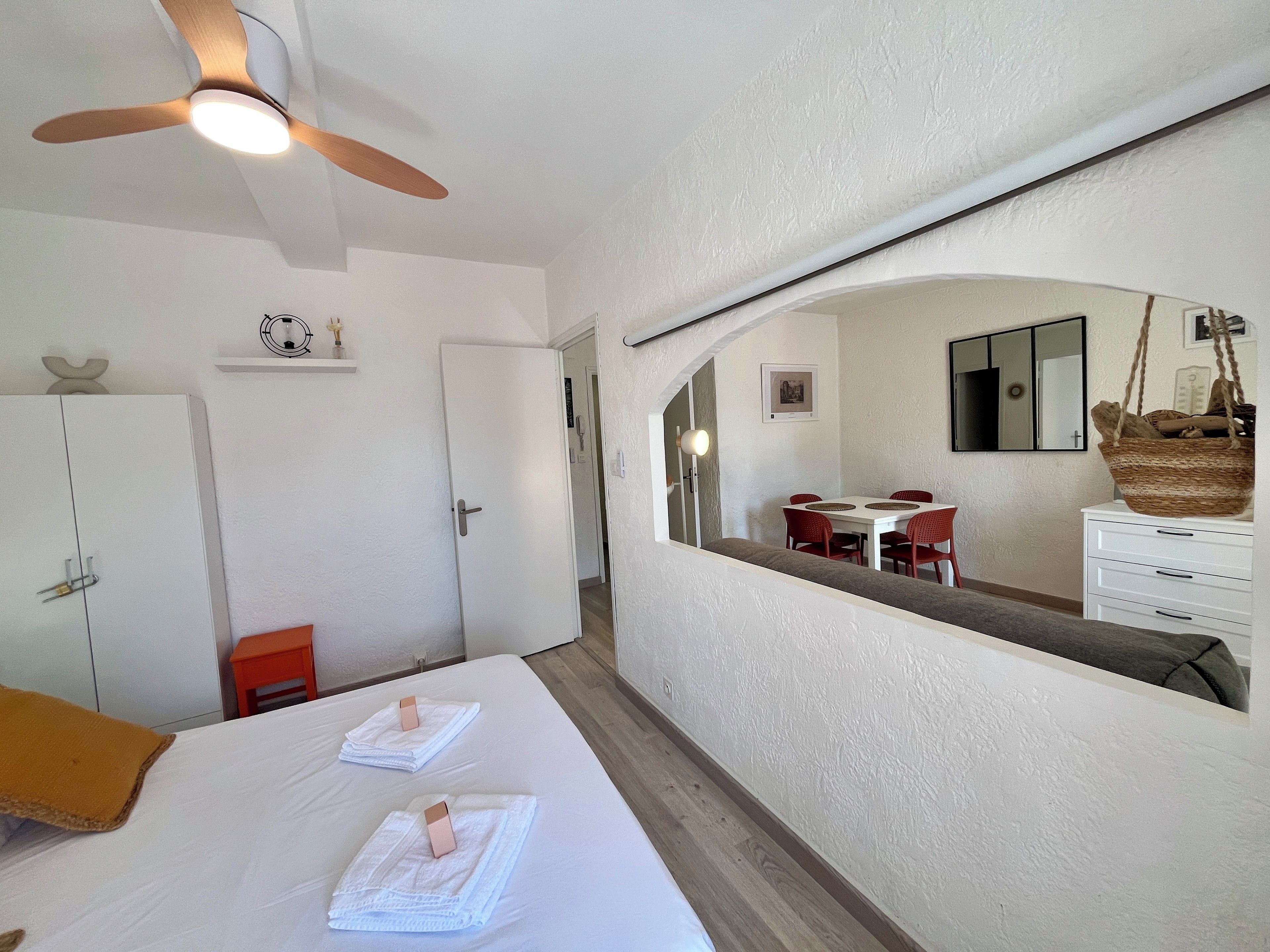 Une chambre à coucher comprenant un lit, un ventilateur de plafond, une armoire et un miroir qui reflète un coin repas.