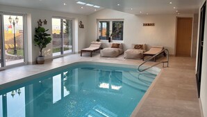 An indoor swimming pool with a clear view of the outside, a wooden ceiling beam, and a seating area with cushions.