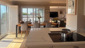 Una cocina moderna con encimera de mármol, comedor con mesa y sillas de madera, y un amplio ventanal con persianas.