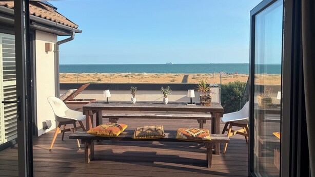 Un balcón con mesa y sillas de madera, vista a la playa y un barco a lo lejos.