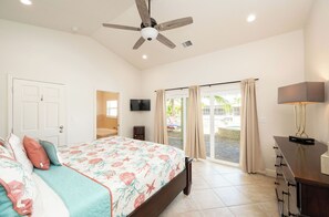 A bedroom with a bed, a ceiling fan, a TV, a desk with drawers, and a sliding glass door leading to an outdoor area.