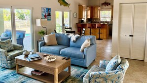 Un salón con un sofá azul, una mesa de centro de madera y un sillón estampado. Se observa una cocina al fondo con armarios de madera y una decoración de pez en la pared.