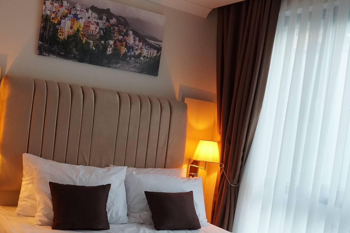 Habitación de hotel con cama, cabecera, un cuadro en la pared, lámpara y ventana con cortinas.