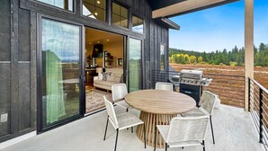 Een modern huis met een terras, compleet met een ronde tafel en stoelen, een barbecue en uitzicht op een bebost gebied.