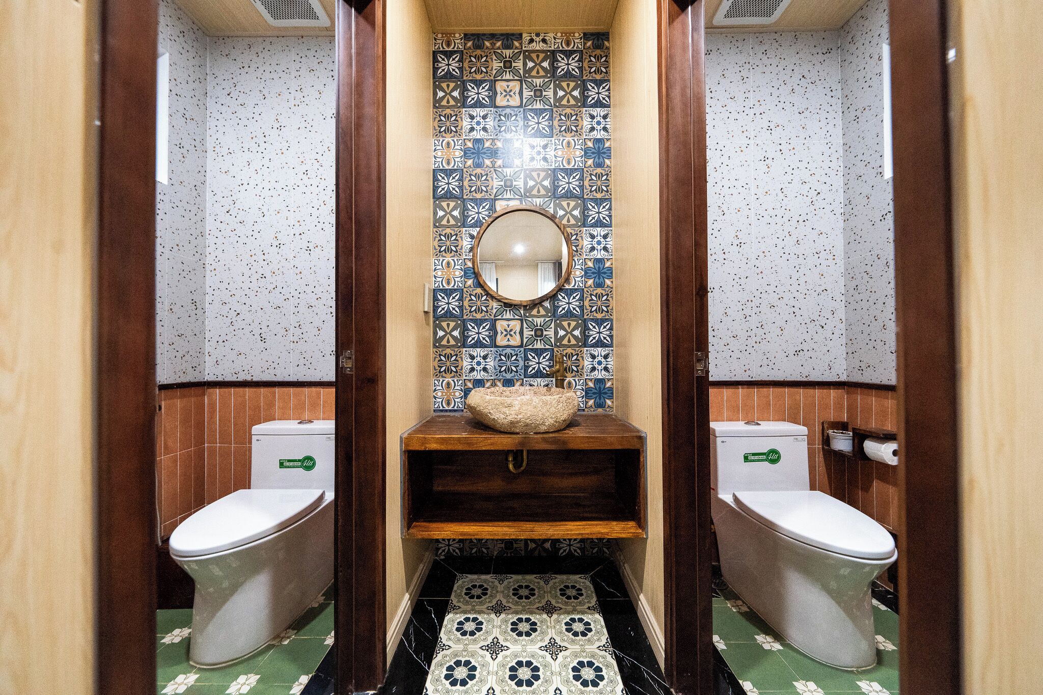 Une salle de bain avec deux toilettes, un lavabo avec un miroir et des carreaux décoratifs aux murs et sur le sol.