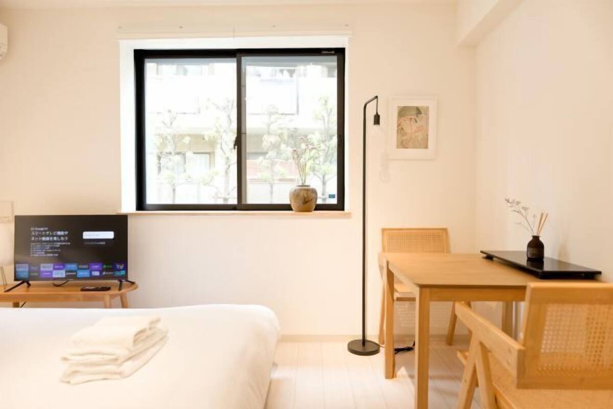Ett hotellrum med en säng, ett skrivbord, en stol, en tv och ett fönster med utsikt över byggnader och träd.