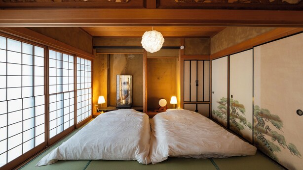 A traditional Japanese room with tatami flooring, a shoji screen wall, a hanging shoji screen, a wooden ceiling with intricate carvings, and a large white bed with two pillows. - Ikkenyahotel FUKUOKA (Fukuoka)