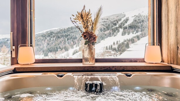 Ein Whirlpool mit Blick auf verschneite Berge durch ein Fenster, eine Vase mit getrockneten Blumen und zwei brennende Kerzen auf der Fensterbank.