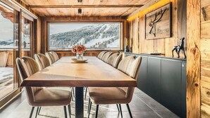 Ein Holzinterieur mit einem großen Esstisch, gepolsterten Stühlen, einer Blumenvase und Blick auf verschneite Berge.