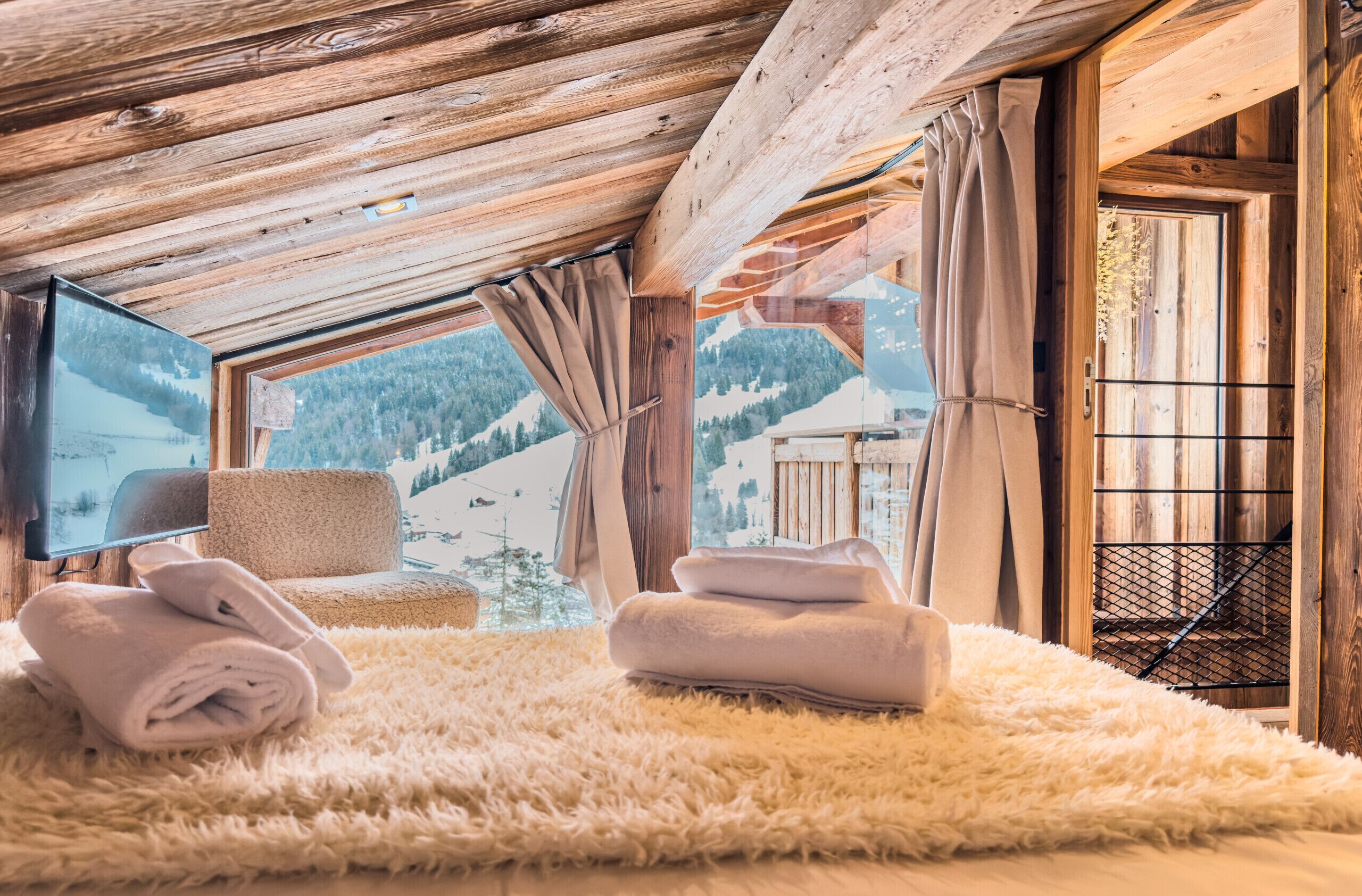 Quarto aconchegante com teto de madeira, uma janela grande com cortinas, tapete felpudo e vista para uma paisagem nevada de montanha.