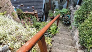 Escadaria de pedra que leva a uma casa com uma pequena janela e um jardim com diversas plantas e guarda-corpo de metal.