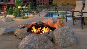 A fire pit surrounded by large rocks, a pool, lounge chairs, and a building in the background.