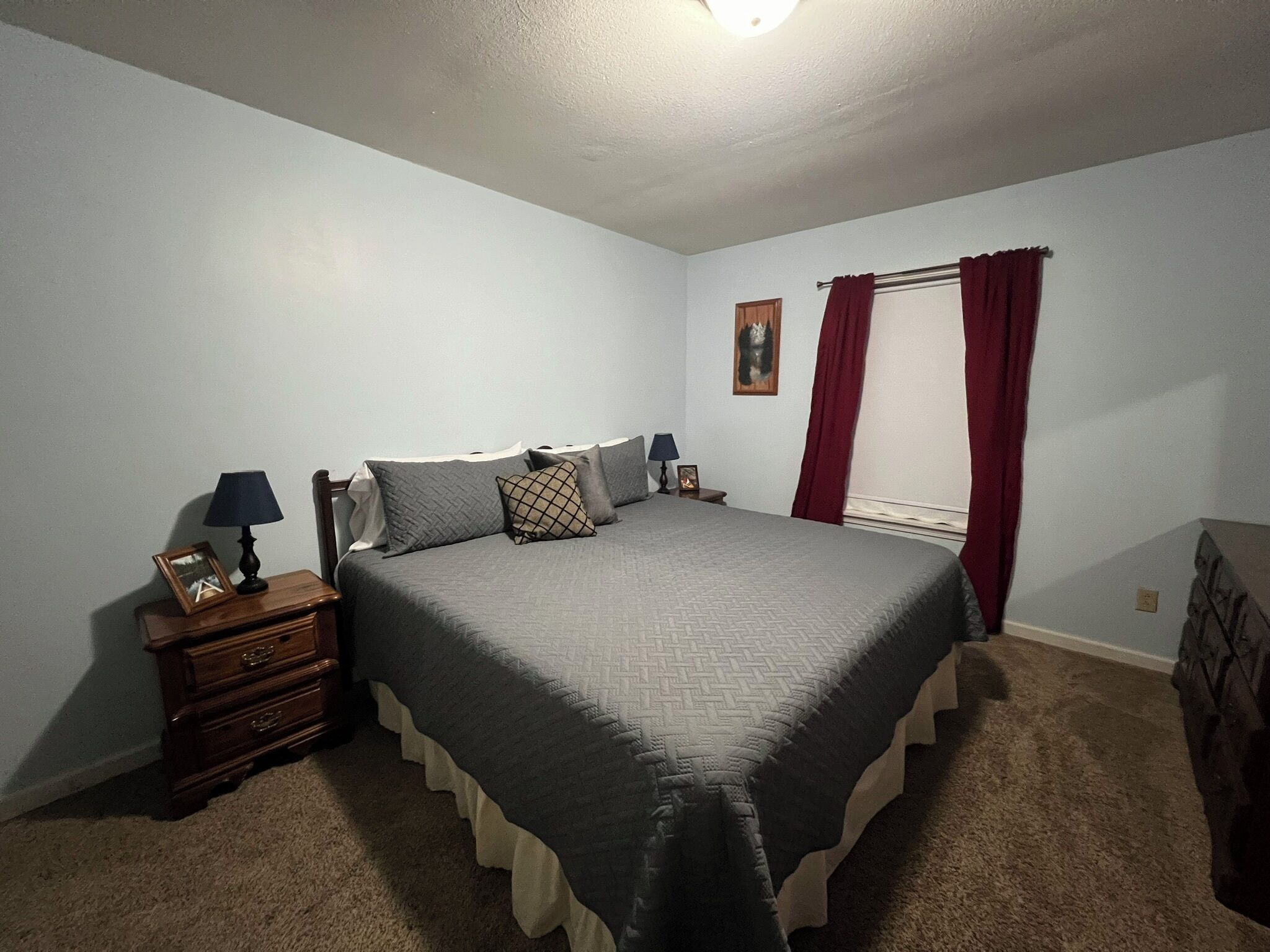 A bedroom with a bed, a nightstand, a dresser, a window with curtains, and a painting on the wall.